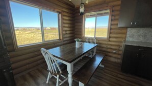 L’intérieur d’un chalet en bois comprend une table à manger, des chaises, un coin cuisine et de grandes fenêtres donnant sur un champ.