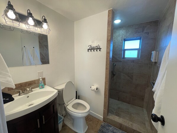 Ein Badezimmer mit Waschbecken, Toilette und einer Duschkabine. Ein Handtuchhalter mit einem Schild befindet sich über der Toilette, und auf dem Waschbecken steht eine Flasche Seife.