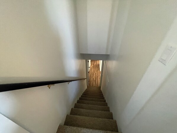 Una escalera estrecha con una puerta de madera en la parte superior, paredes blancas y una barandilla negra.