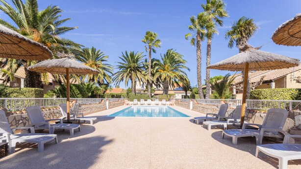 Un espace piscine agrémenté de chaises longues et de parasols en chaume, entouré de palmiers et surplombé d’un ciel dégagé.