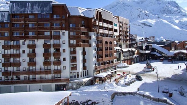 Un hotel de varios pisos con balcones, una pista de esquí y montañas nevadas al fondo.