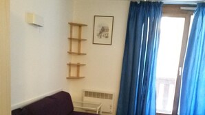 Una habitación con un sofá morado, una mesita, un estante en la pared, un cuadro enmarcado y una ventana con cortinas azules.