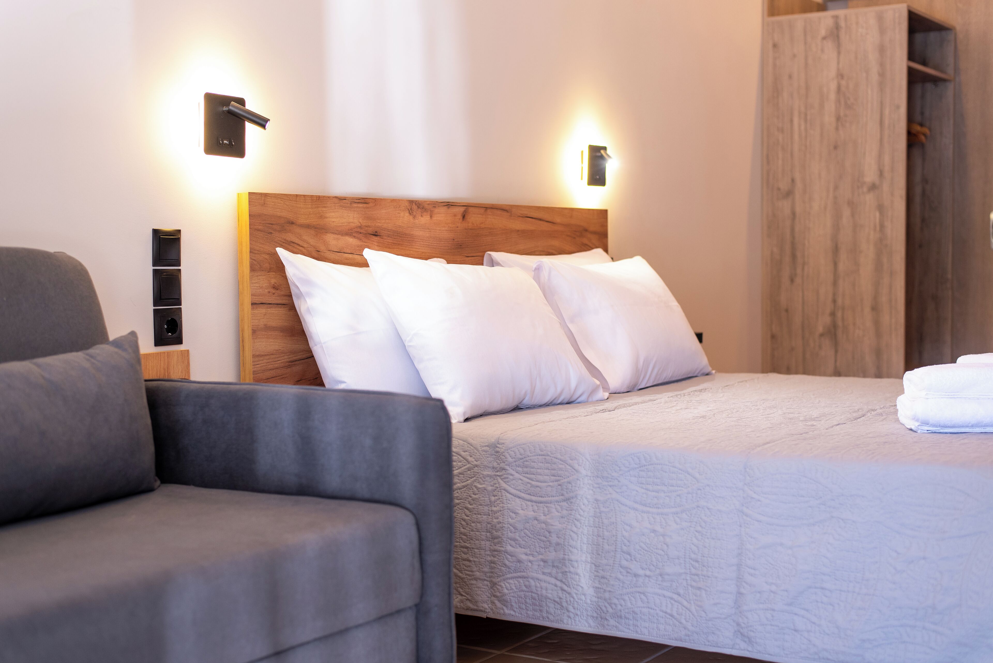 A modern bedroom with a wooden headboard, white bedding, and a grey sofa.