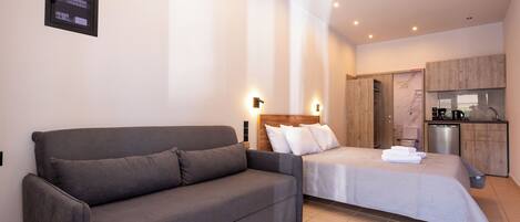 A modern bedroom with a grey sofa, a bed with white bedding, a wooden headboard, and a small kitchenette in the background.