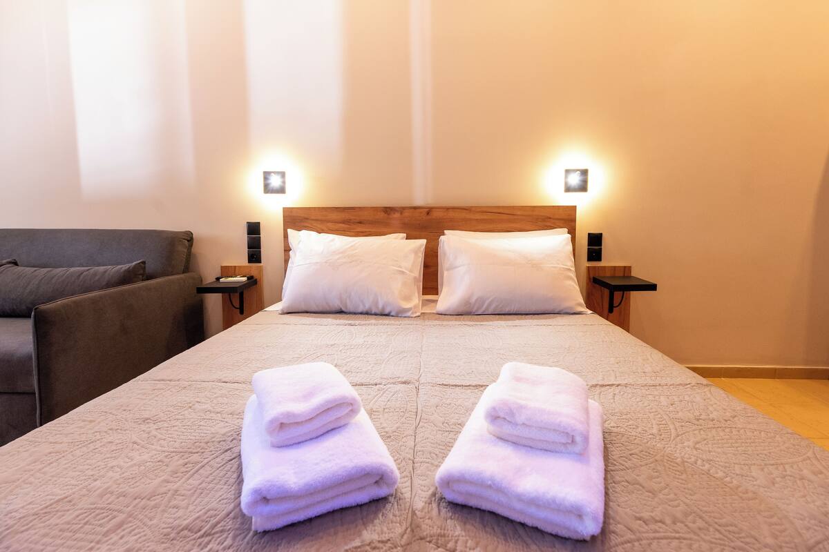 A neatly made bed with two pillows, a bedside table with a lamp, and a couch in a hotel room.