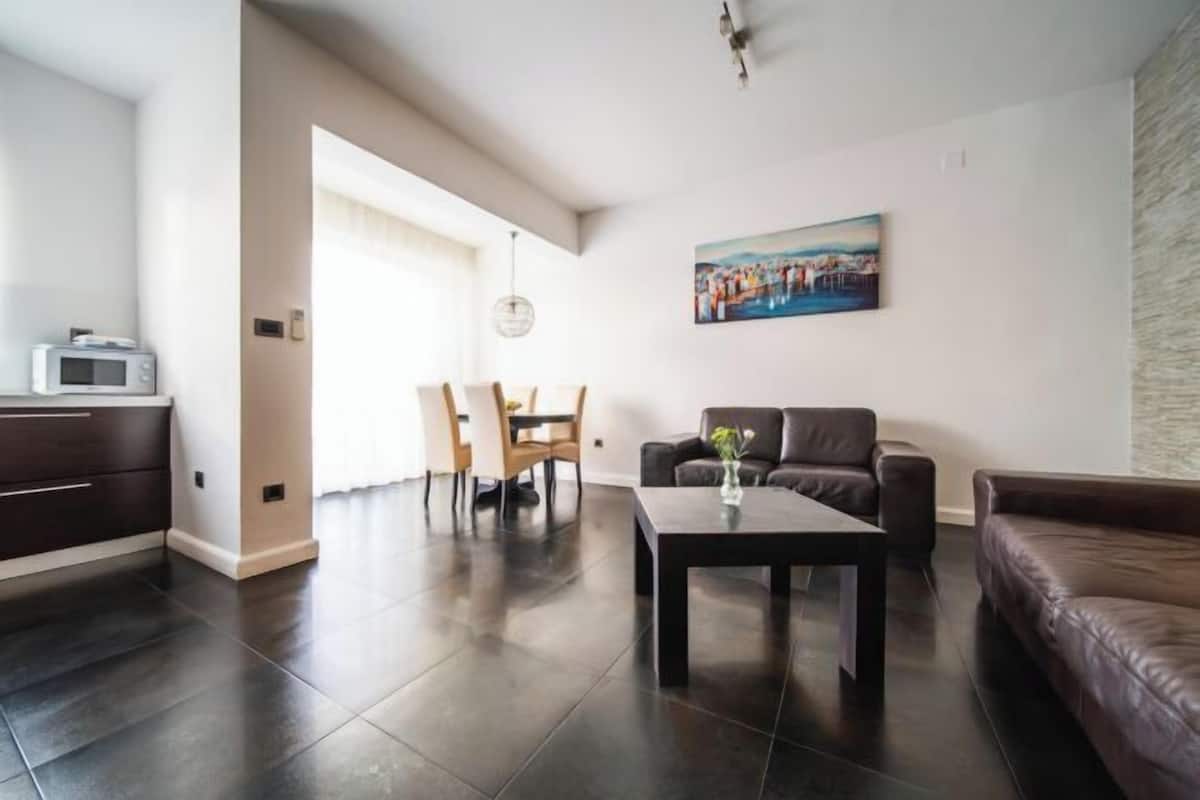 Una sala moderna con piso de baldosas oscuras, un sofá de cuero marrón, un comedor con sillas de madera y un cuadro en la pared.