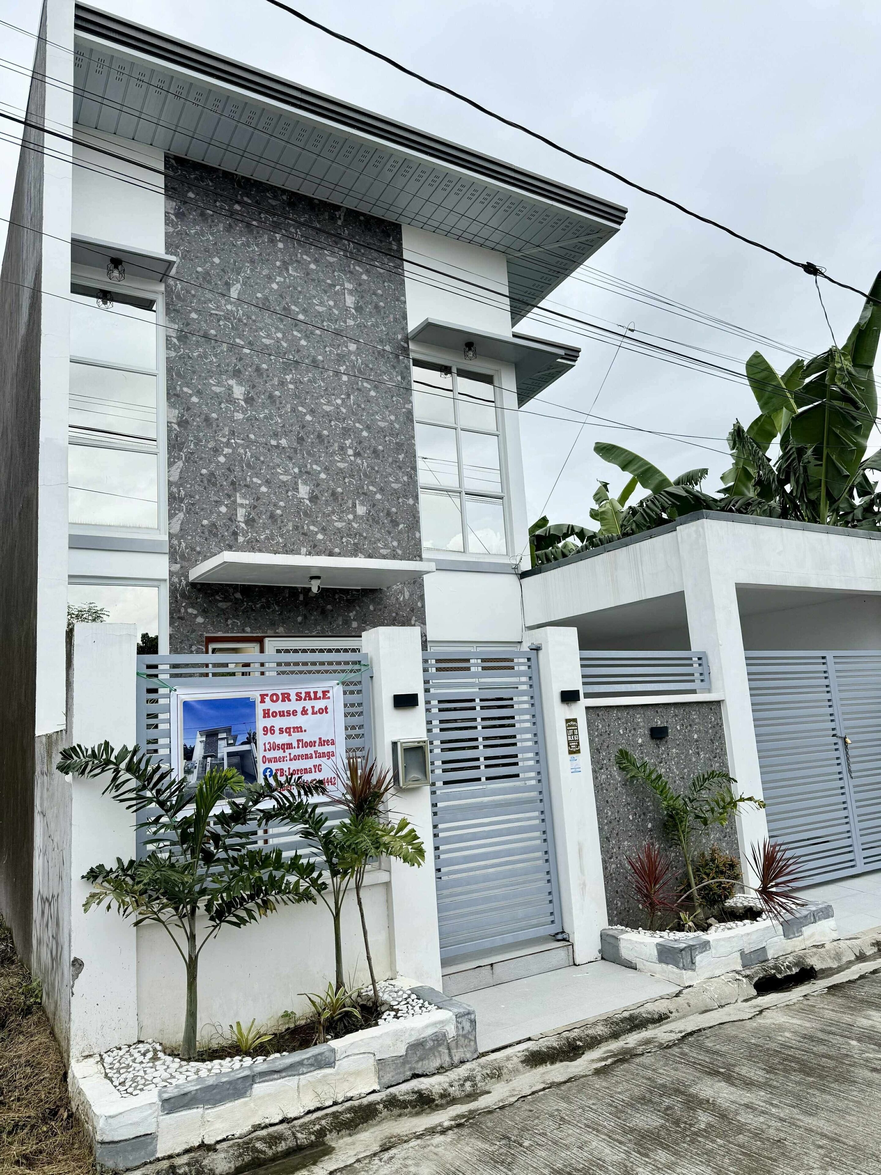 Una casa moderna de dos pisos con un letrero de 'En venta', entrada protegida por una reja y un pequeño jardín.