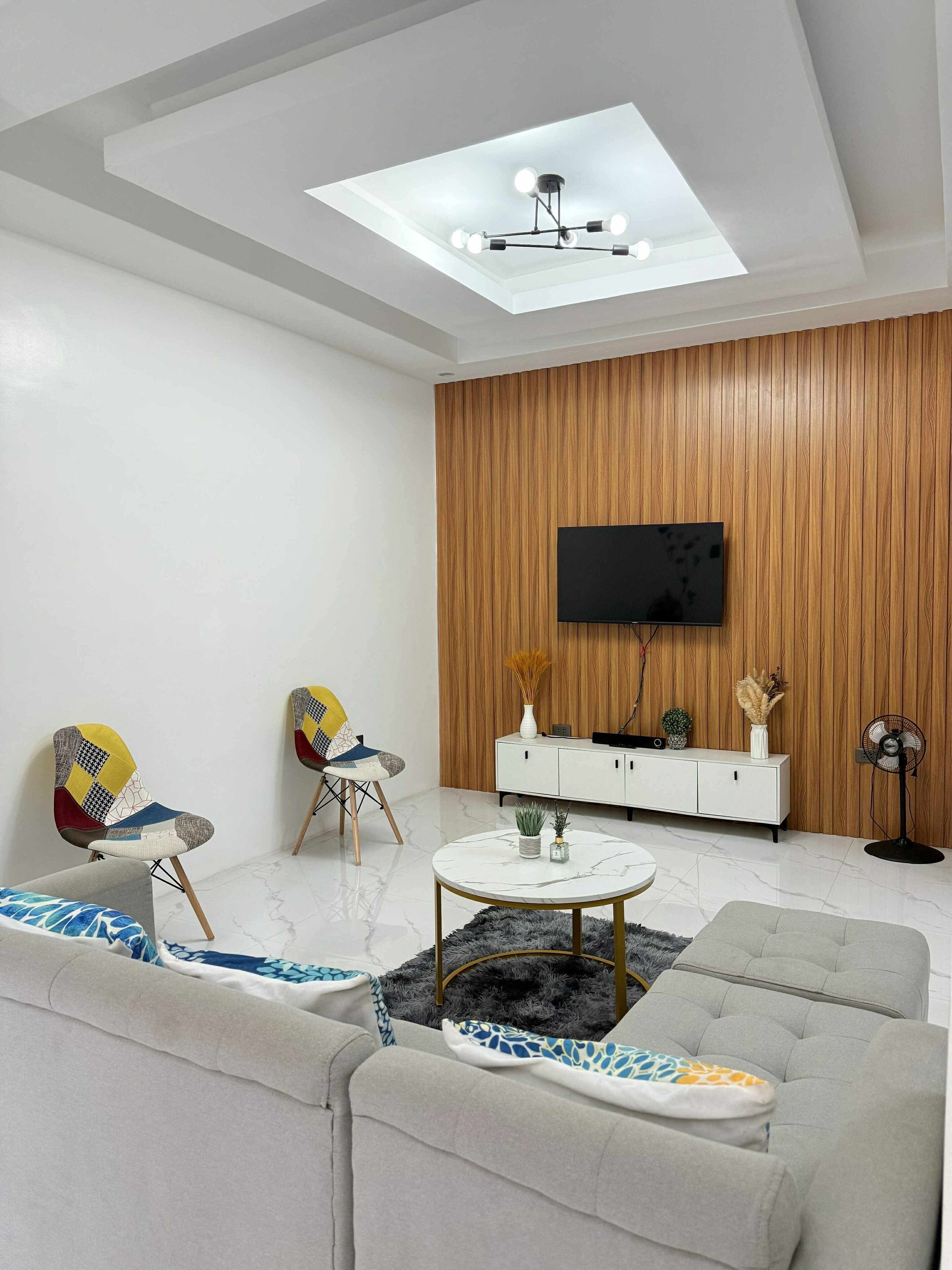 A modern living room with a sofa, two chairs, a coffee table, a TV mounted on a wooden wall, and a ceiling light fixture.