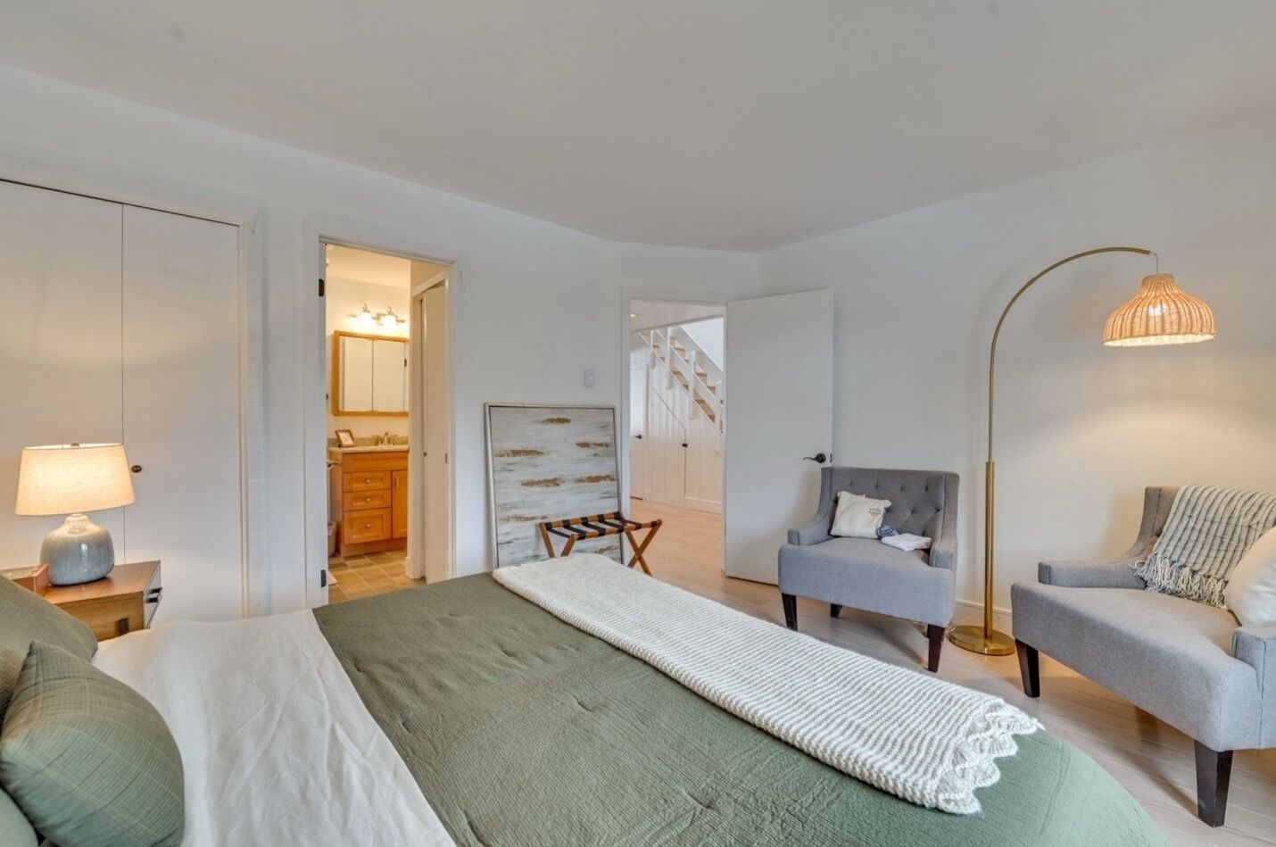 Ein modernes Schlafzimmer mit einem Bett, zwei Sesseln, einer Stehlampe und Blick auf eine Treppe.
