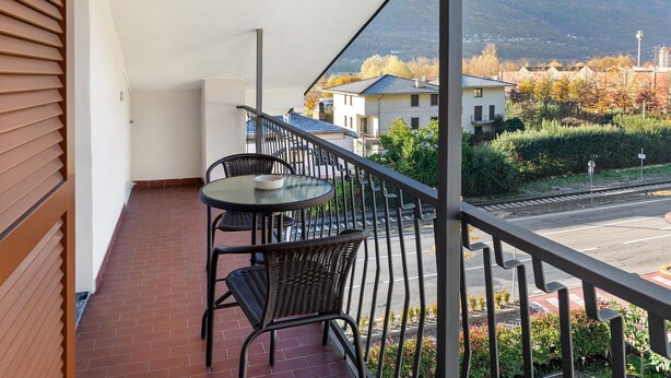 Varanda com mesa e cadeiras, vista para uma rua e casas ao fundo.