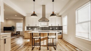 Una cocina moderna con piso de madera, dos lámparas colgantes, una isla central con taburetes de bar y un ventanal con persianas.