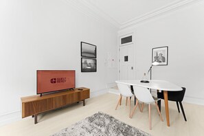 Ein modernes Hotelzimmer mit einem Holzschrank, einem Flachbildfernseher, einem weißen Esstisch mit Stühlen und einem grauen Teppich.