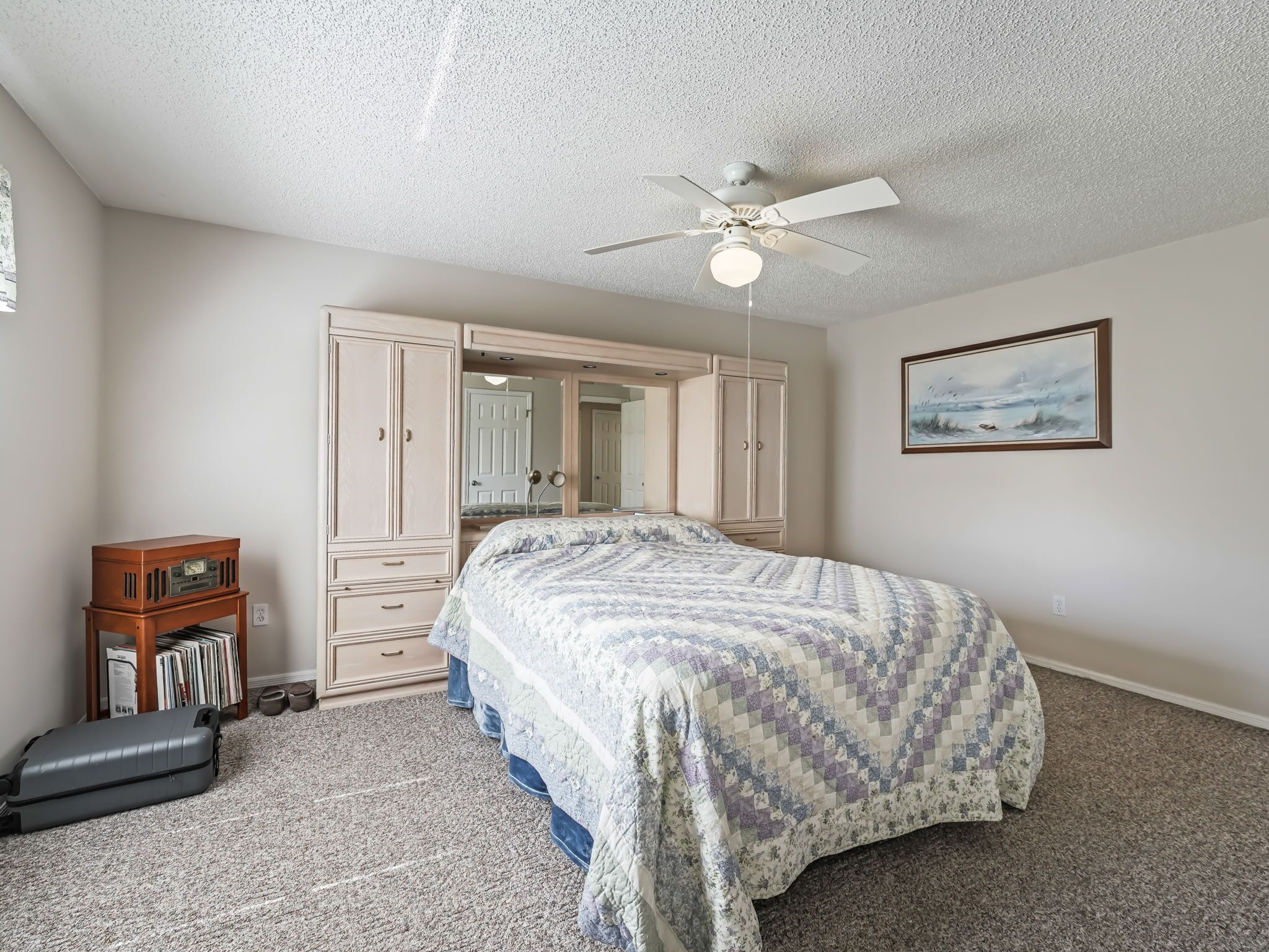 A bedroom with a bed, a wardrobe, a ceiling fan, a bookshelf, and a painting on the wall.