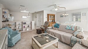 A living room with a sofa, a coffee table, a blue armchair, and a wooden cabinet. - 2/2 Patio Villa in De La Vista (The Villages)
