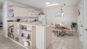 A compact kitchen with a microwave, stove, and refrigerator. There is a dining area with a table and chairs, and a bookshelf filled with books and decorative items. - 2/2 Patio Villa in De La Vista (The Villages)
