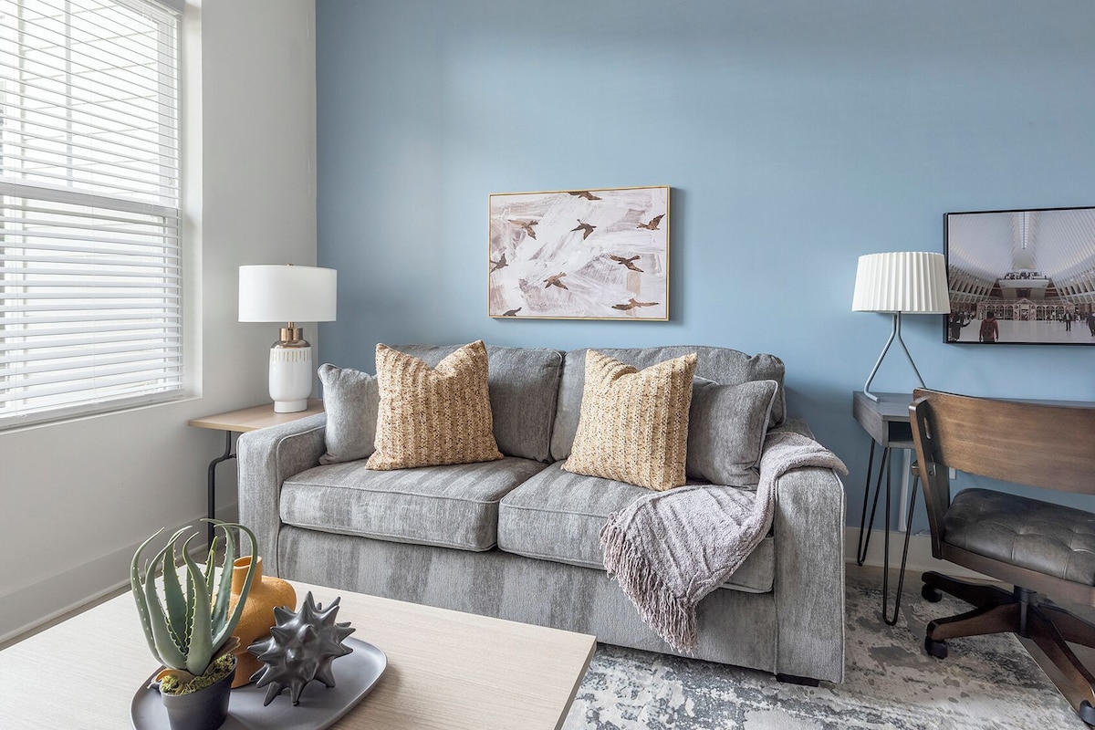 A living room with a grey sofa, two decorative pillows, a table with a plant, a lamp, and a framed picture on the wall.