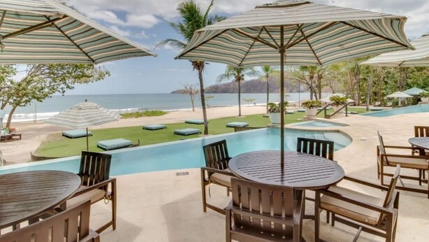 Un espace piscine doté de chaises longues et de parasols, avec vue sur une plage et l’océan.