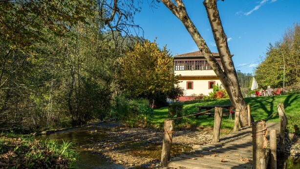A wooden bridge over a stream, surrounded by trees and a building with a red roof.