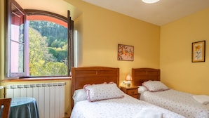 A bedroom with two beds, a window with an open shutter, a table, and a radiator.