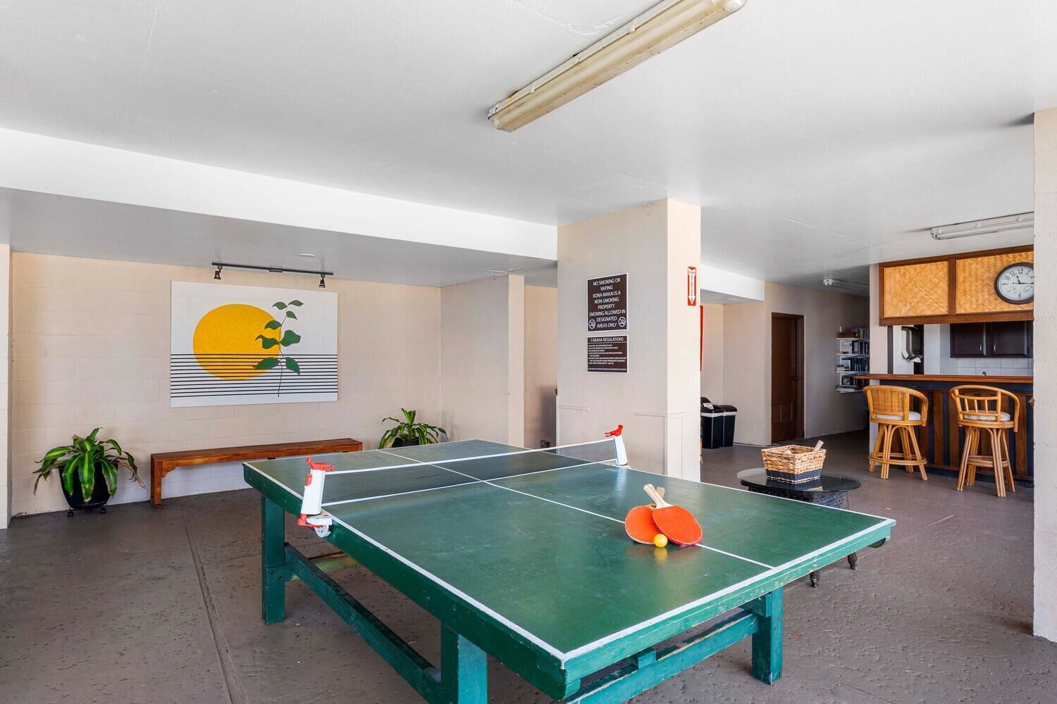 Une table de ping-pong dans une pièce qui comprend un coin cuisine, un banc et une horloge fixée au mur.
