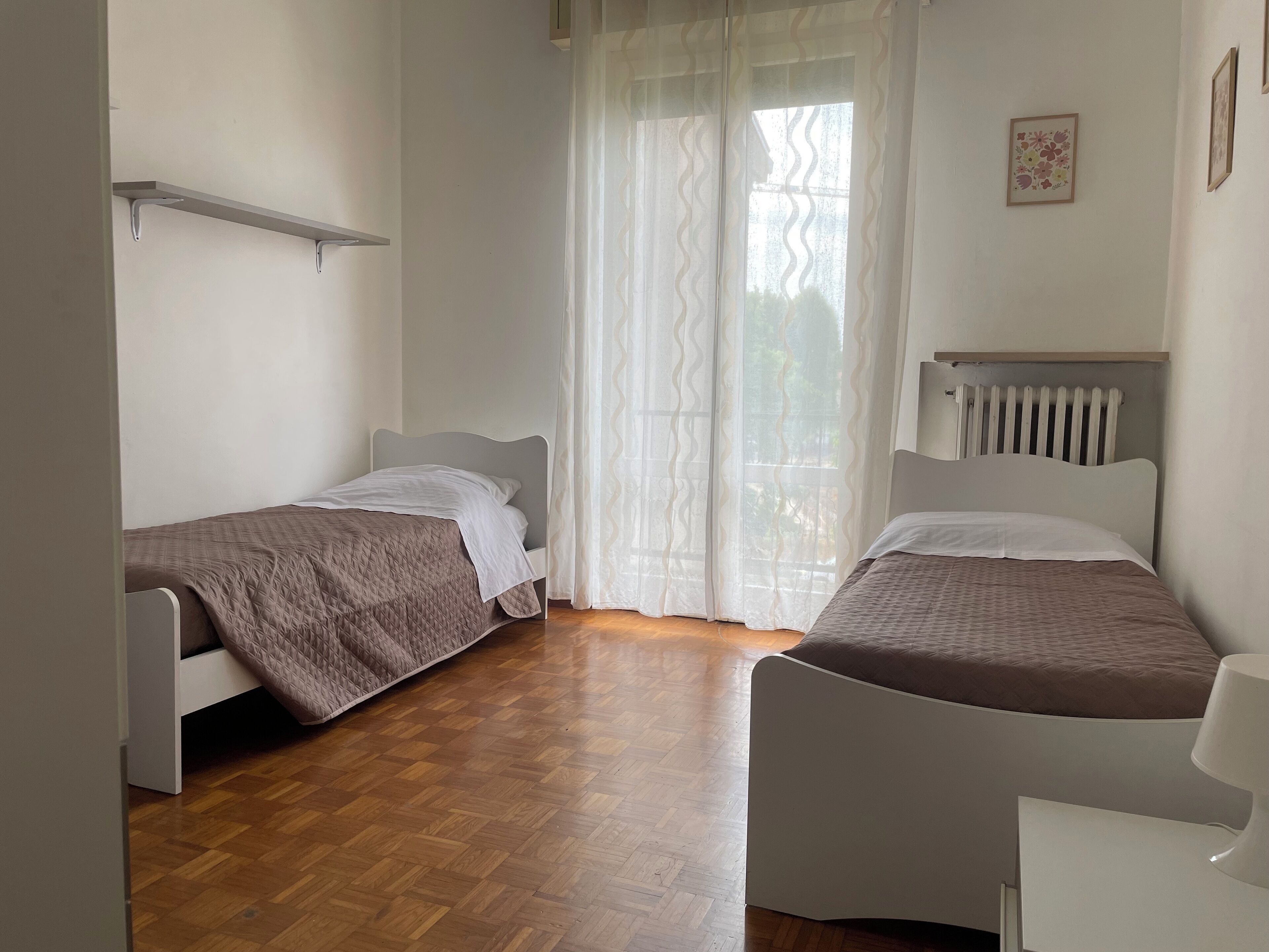Una camera da letto con due letti singoli, pavimento in legno, un termosifone e una finestra con tende leggere.