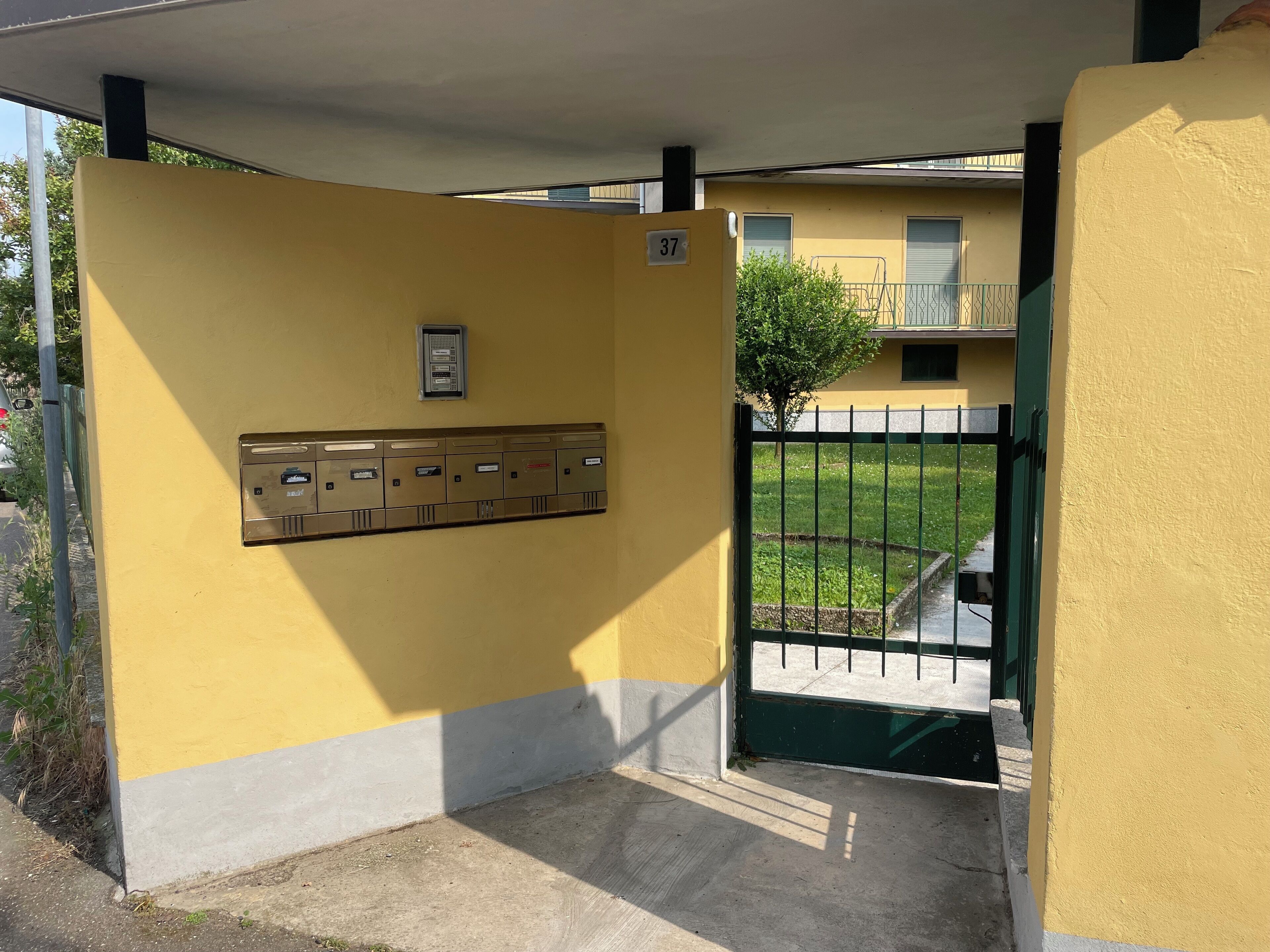 A yellow building with a gated entrance, a mailbox, and a small tree in the background.