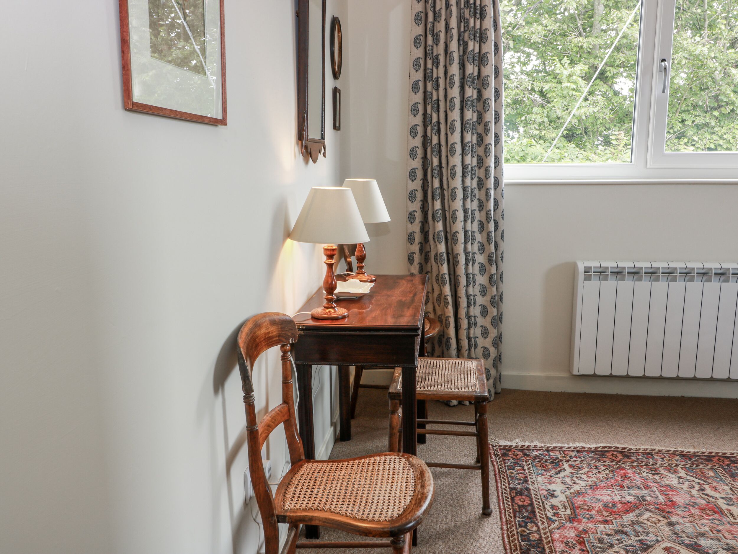 Una habitación con una mesa de madera, dos sillas, una lámpara, un radiador y una ventana con cortinas.