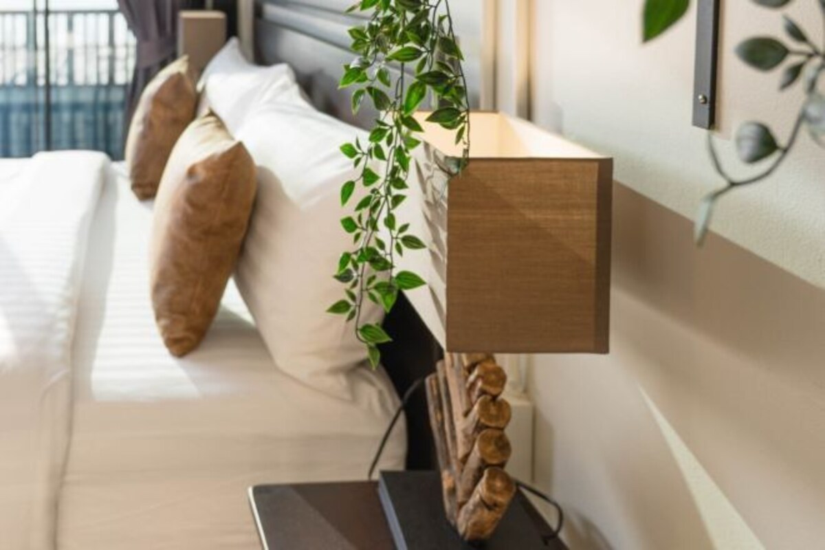 A neatly made bed with white linens and two brown pillows, a wooden bedside table with a plant, and a wall-mounted light fixture.