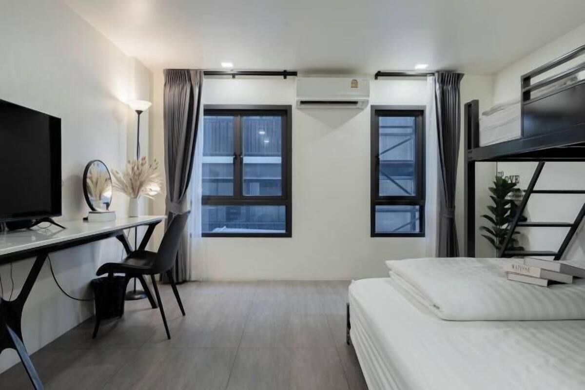 Una habitación de hotel pequeña con una cama, un escritorio con silla, un televisor y dos ventanas con cortinas.