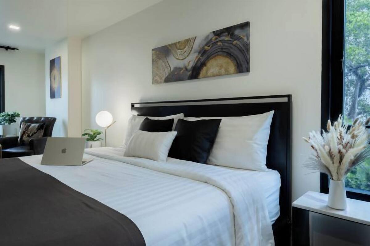 Una habitación de hotel moderna con una cama grande, una computadora portátil sobre la cama, una mesita de noche con una lámpara y una obra de arte en la pared.