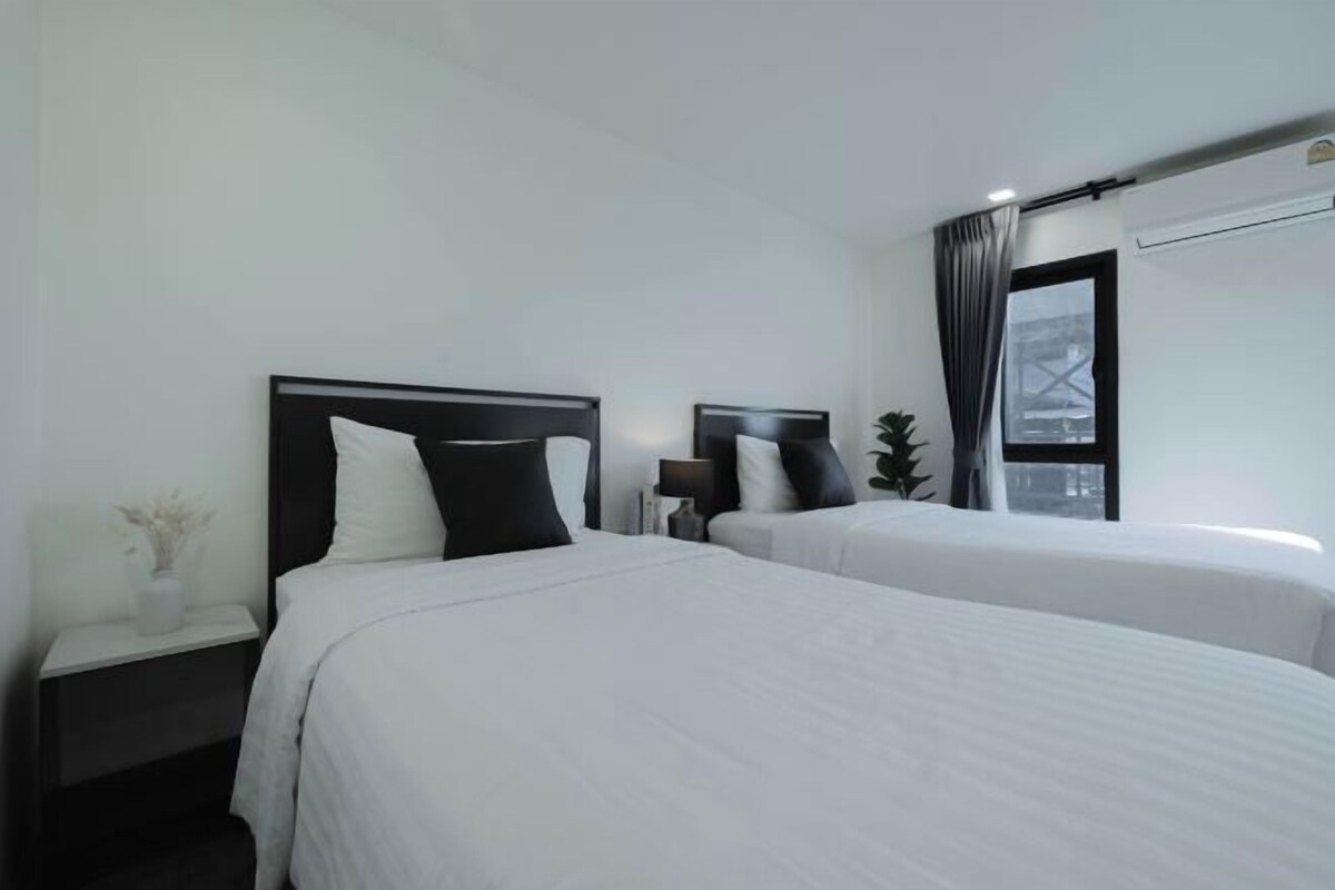 Habitación de hotel con dos camas individuales, una mesita de noche con lámpara, una planta pequeña y una ventana con cortinas.