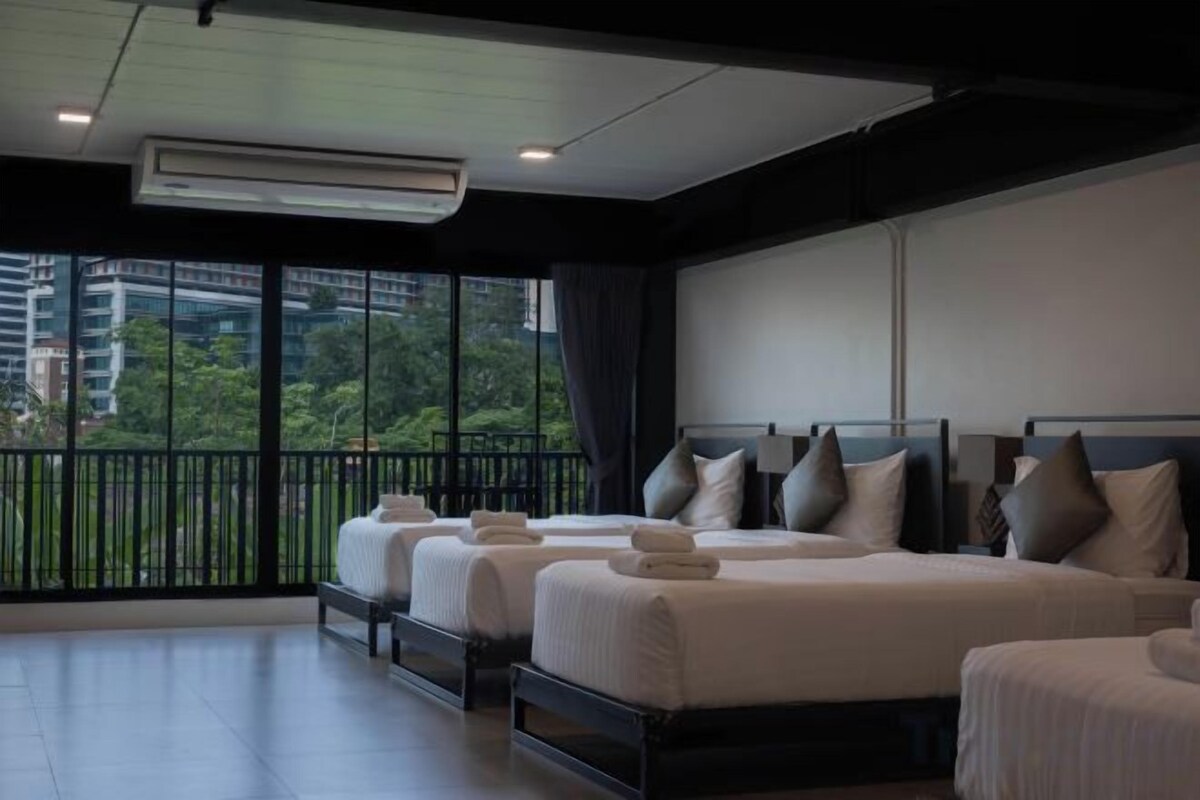 A hotel room with three beds, a balcony with a view of buildings and trees, and an air conditioning unit.