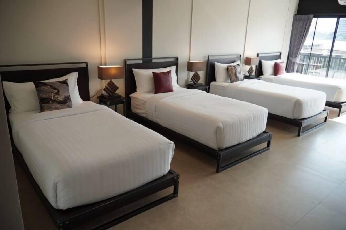 Habitación de hotel con tres camas individuales, cada una con estructura de madera y ropa de cama blanca. Hay mesitas de noche con lámparas, y la habitación tiene un ventanal que ofrece vistas al exterior.