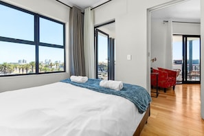 Une chambre d’hôtel avec un grand lit, une vue sur les palmiers et les immeubles, et un balcon avec des chaises rouges.