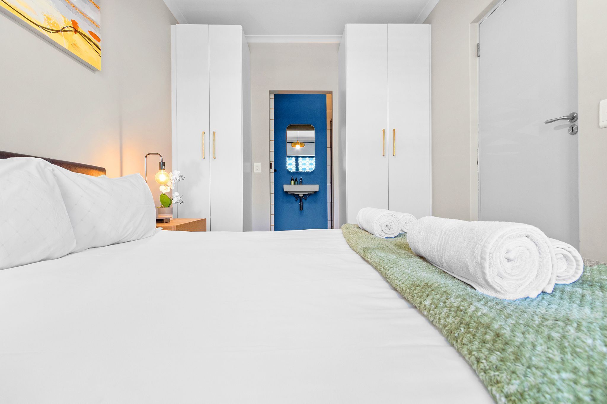 A hotel room with a bed, a nightstand, a blue door, and a bathroom visible through the door.