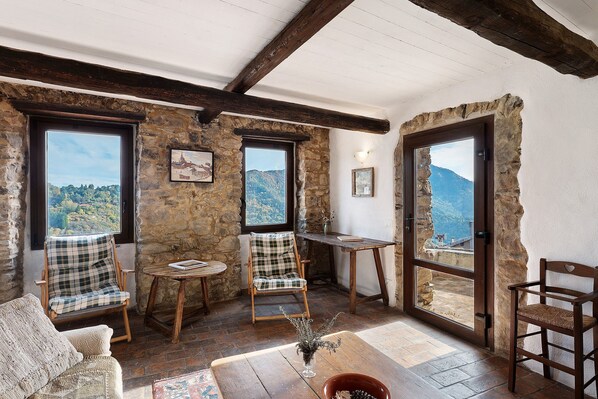 A rustic room with stone walls, wooden beams, a view of the mountains, and a cozy seating area with a table and chairs.