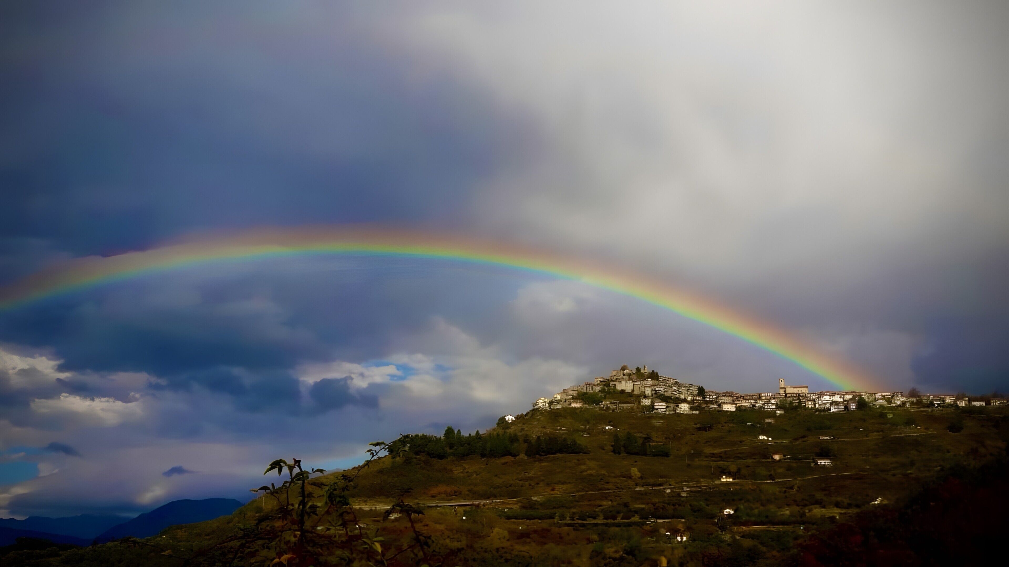 Paisatge amb un arc de l'iris que voreja un turó on hi ha edificis.