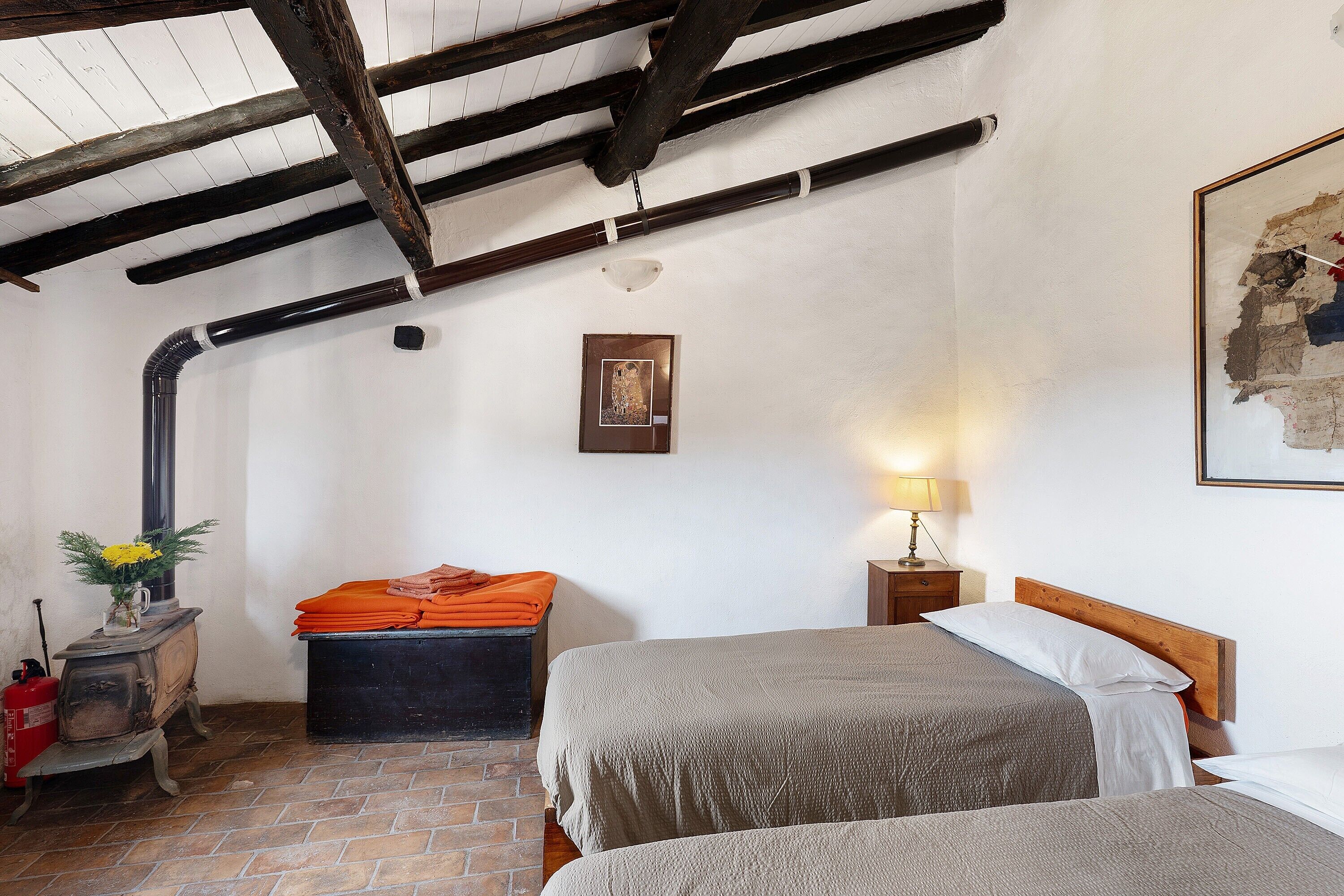 A room with two beds, a wooden ceiling, a brick floor, a small table with a lamp, and a heater.