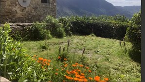 A stone building with a window, surrounded by greenery and a mountainous backdrop.
