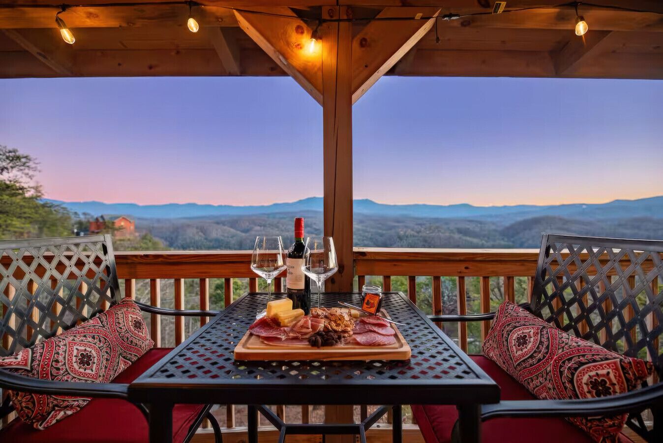 A covered outdoor seating area with a table set for a meal, featuring a bottle of wine, glasses, and a variety of meats.