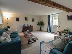 Ein Wohnzimmer mit Türkis-Sofas, einem Holztisch mit Büchern, einem kleinen Beistelltisch mit Lampe und einem großen Fenster mit Vorhängen.