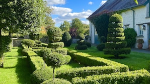 A well-manicured garden with neatly trimmed hedges and a variety of trees, leading to a white building with a stone facade. - West Cottage (Llandovery)
