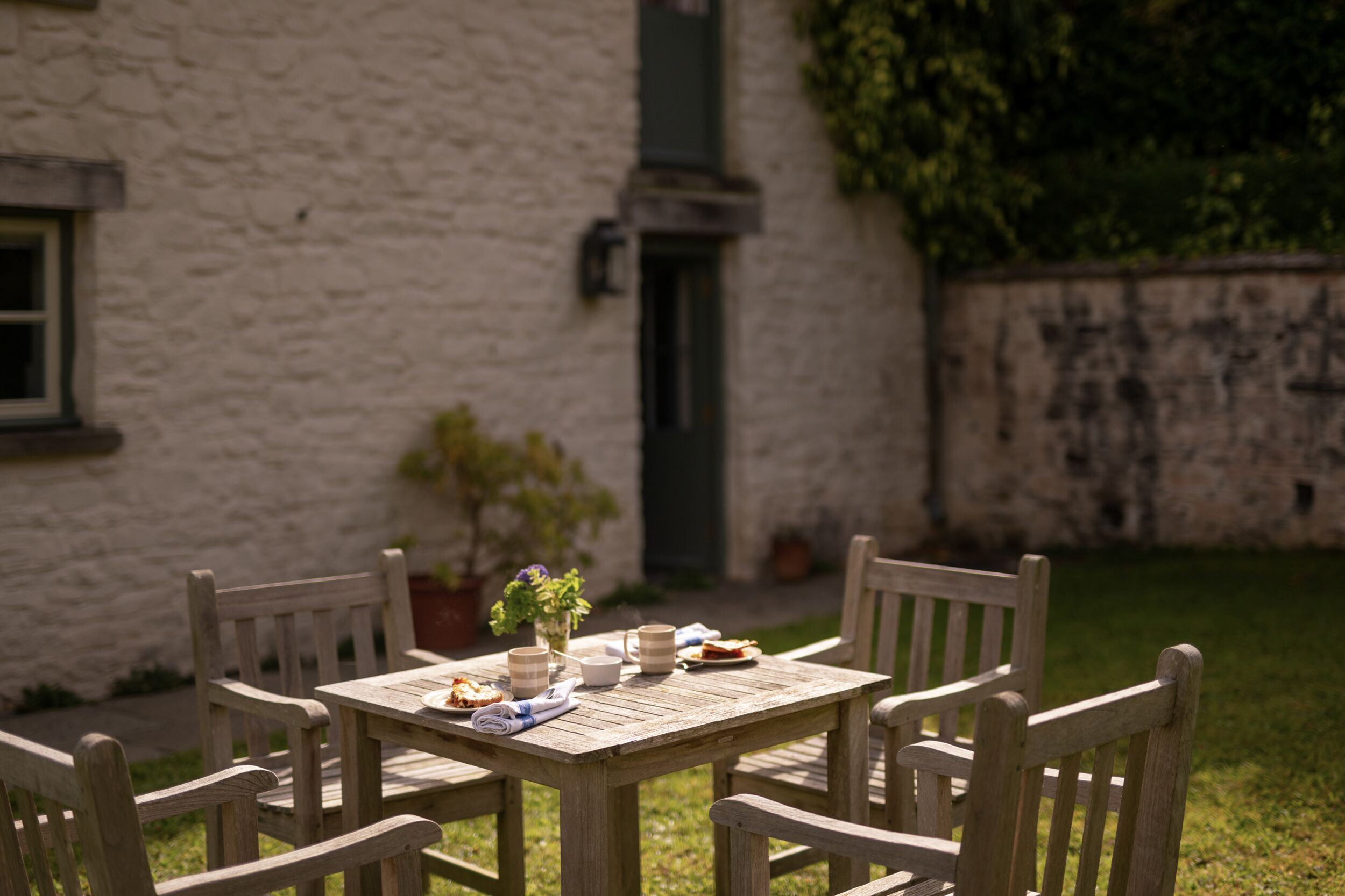 A wooden table with chairs, a vase with flowers, and a plate of food.