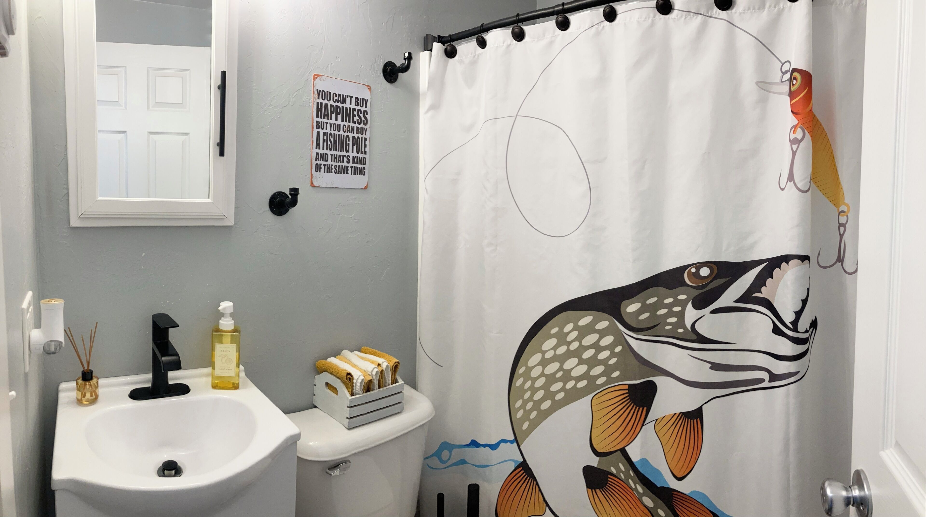 Et badeværelse med et hvidt håndvask, toilet, bruseforhæng med fiskemotiv, et skilt på væggen og et vindue med hvid ramme.