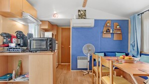 A compact kitchenette with a microwave, toaster, and coffee maker, a dining area with a table and chairs, and a blue accent wall with shelves. - Mobile Home for 6 with Private Terrace, Wi-Fi & Air Conditioning in Hyères (Hyères)