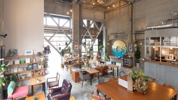 Un café spacieux avec de hauts plafonds, des poutres apparentes et de grandes fenêtres qui laissent entrer la lumière naturelle. On y trouve plusieurs espaces aménagés avec des tables et des chaises, ainsi que des étagères remplies de livres et d’objets décoratifs.