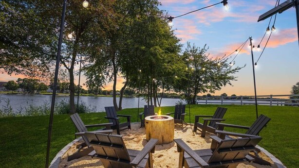 A circular outdoor seating area with a central fire pit surrounded by chairs, set against a backdrop of a lake and trees.