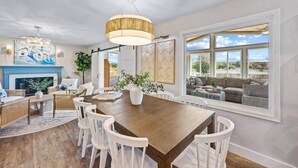 A spacious dining area with a wooden table and white chairs, a fireplace, and large windows offering a view of the outdoors.
