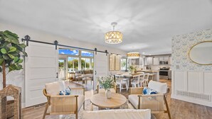 A spacious living area with a large dining table, comfortable seating, a kitchen in the background, and a view through a sliding door.