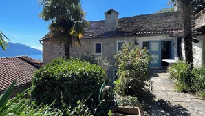 A stone house with a tiled roof, a small garden with various plants, and a view of the sea. - Casa del Prete (Cannero Riviera)